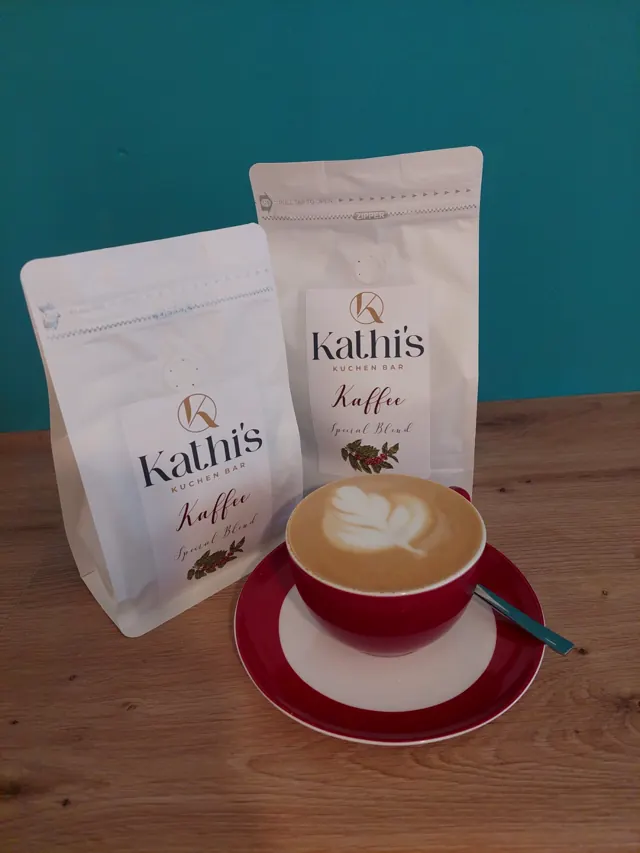 Cappuccino mit Latte Art vor zwei Tüten Kathi's Kaffee Special Blend