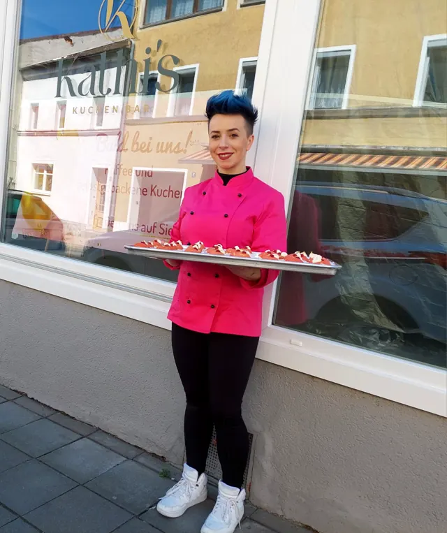 Kathi in pinker Kochjacke vor dem Schaufenster der Kuchenbar, mit einem Tablett voller Törtchen