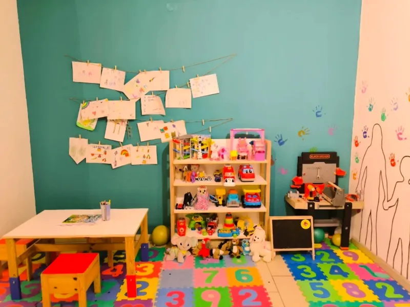 Kinderecke vor der türkisen Wand: Maltisch mit Hocker, Spielzeugregal, Kinderwerkbank, Tafel und bunte Puzzlematte, an der Wand hängen Kinderzeichnungen